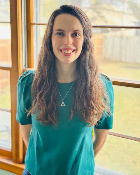 Amanda O’Connell smiling, standing by a window with wooden frames and trees behind her.